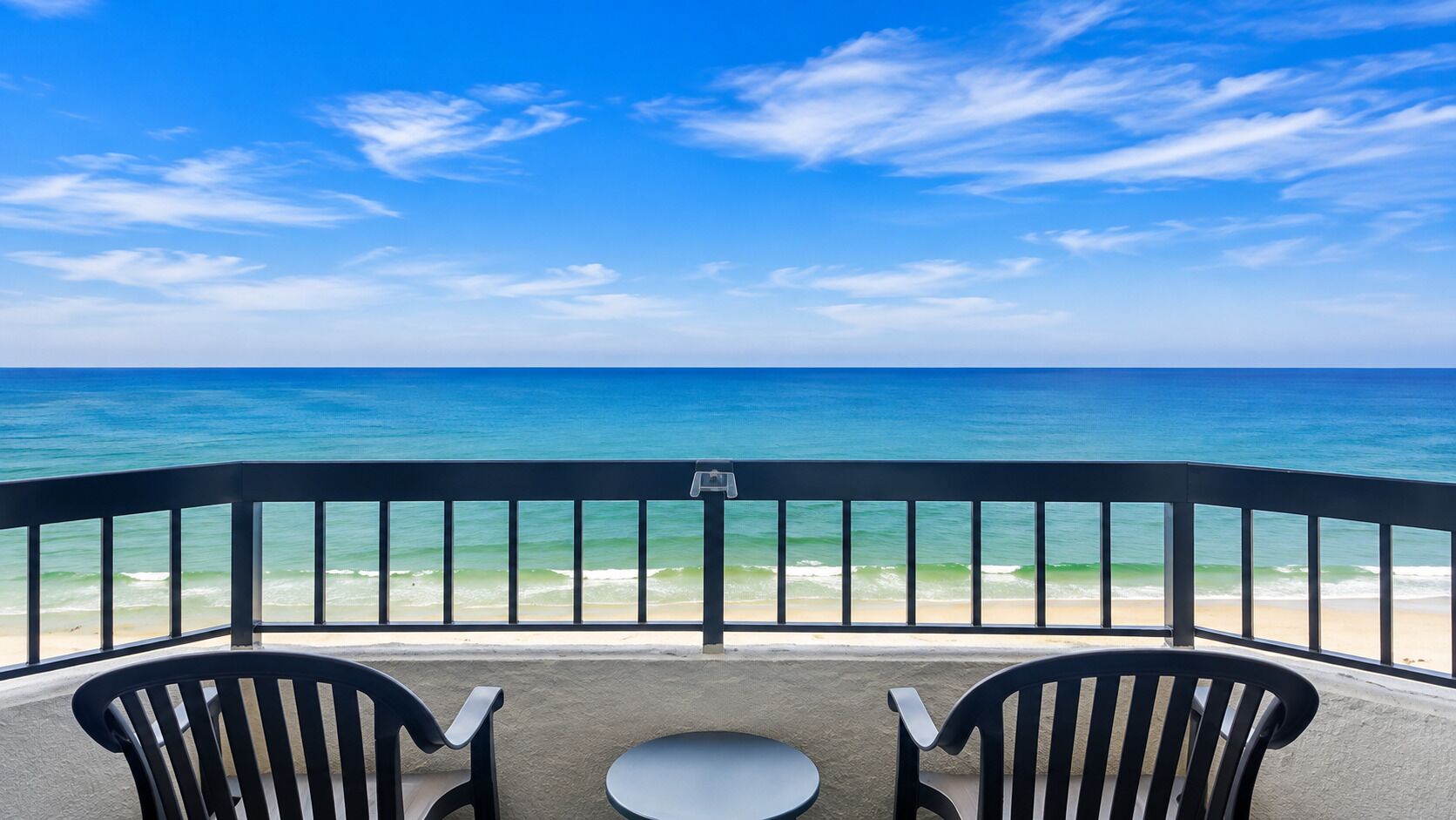 A balcony with two chairs and a small table, overlooking a beach with waves and a clear sky.