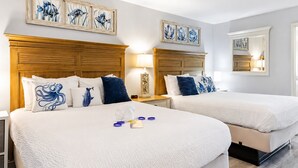Ein Hotelzimmer mit zwei Betten, jeweils mit weißer Bettwäsche und blauen Zierkissen. An den Wänden über den Betten befinden sich Wandbilder und ein Spiegel.