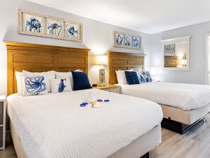A hotel room with two beds, each with white bedding and blue decorative pillows. There are wall-mounted paintings above the beds and a mirror on the wall. - The Peachy Palm Oceanfront at Compass Cove (Myrtle Beach)