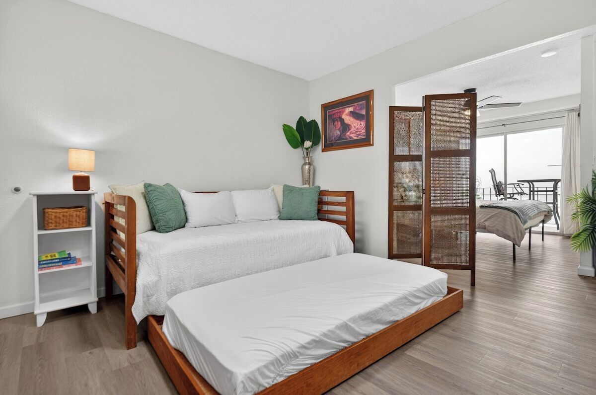 A bedroom with a large bed, a nightstand, a bookshelf, a plant, and a sliding door leading to a balcony.