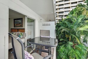 Ein Balkon mit Glastisch und Stühlen, eine Schiebetür zu einem Schlafzimmer und Blick auf ein hohes Gebäude sowie dichtes Grün.