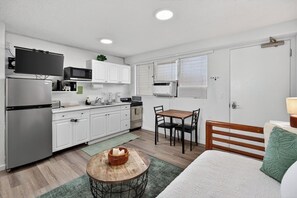 Eine kompakte Küche mit modernen Geräten, ein Essbereich mit Holztisch und ein Bett mit einem grünen Kissen.