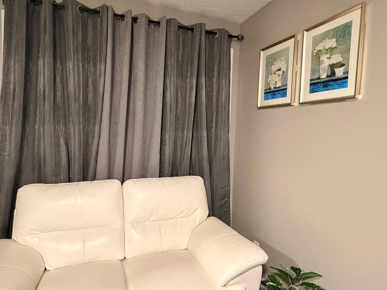 Un canapé beige dans une pièce où l’on aperçoit des rideaux gris et deux tableaux encadrés accrochés au mur.