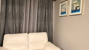 Un canapé beige dans une pièce où l’on aperçoit des rideaux gris et deux tableaux encadrés accrochés au mur.