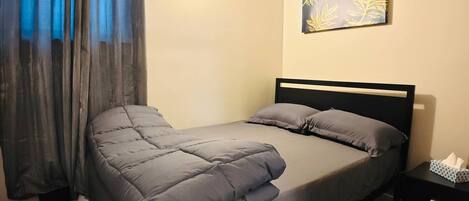 A single bed with a black headboard, grey bedding, a bedside table with a tissue box, and a wall-mounted artwork.