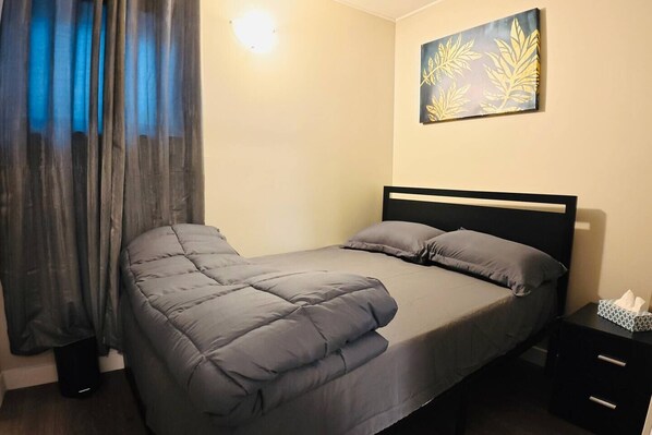 A single bed with a black headboard, grey bedding, a bedside table with a tissue box, and a wall-mounted artwork.
