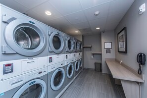 A laundry facility with multiple washing machines, a trash bin, a phone, and a framed notice on the wall.