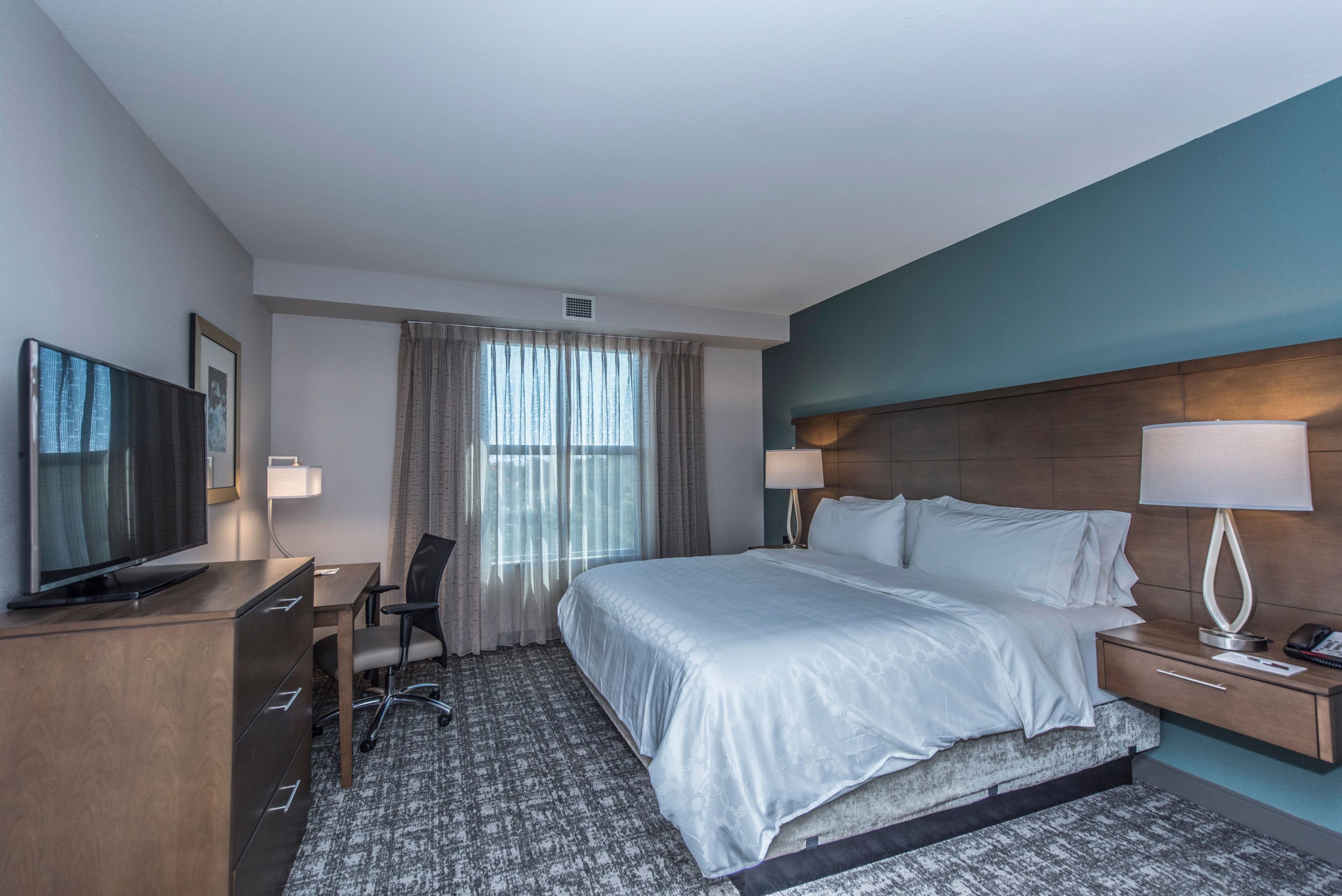 Une chambre d’hôtel avec un lit, un bureau avec une télévision, une chaise, une lampe et une fenêtre avec des rideaux.
