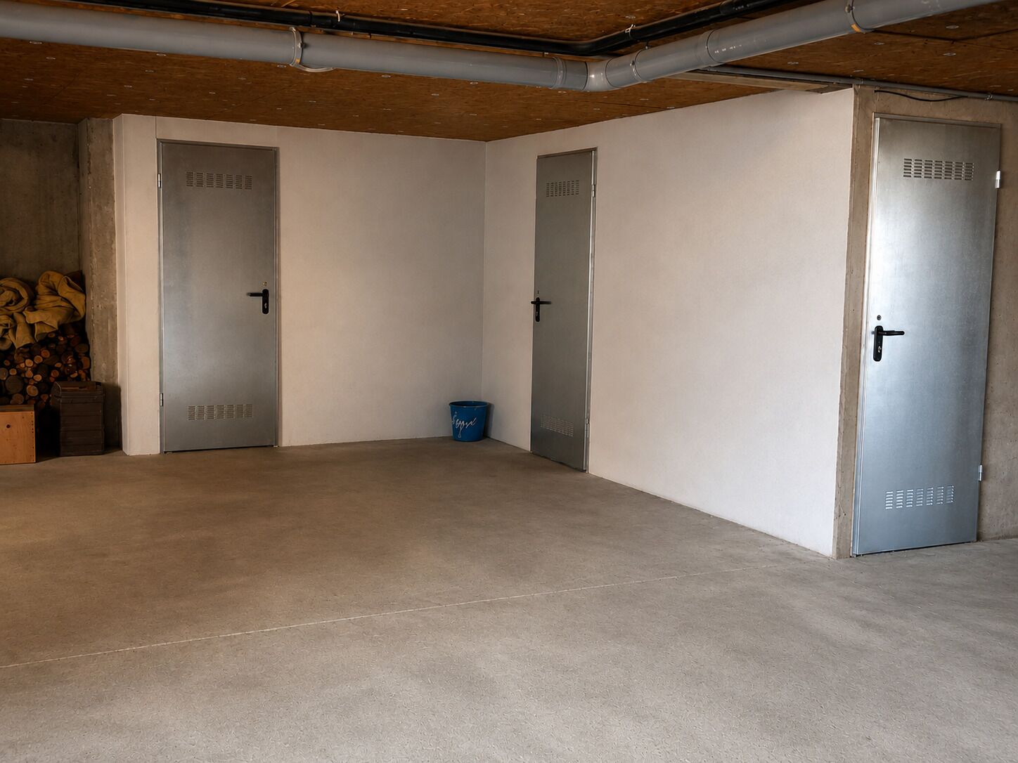A spacious, empty room with a concrete floor, three metal doors, and a blue bucket on the floor.