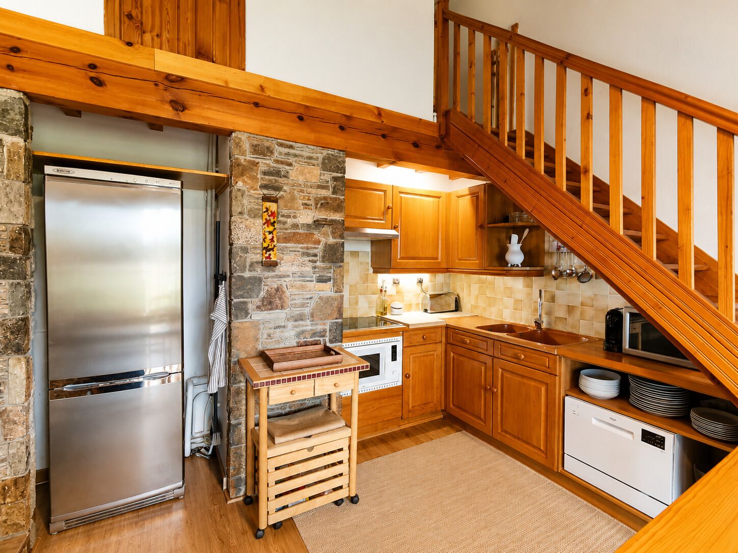A kitchen with a stone wall, wooden cabinets, a stainless steel refrigerator, a wooden cart, and a wooden staircase.