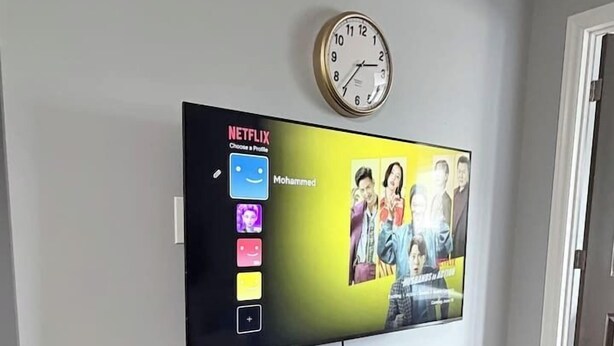A flat-screen TV mounted on a wall with a Netflix interface, a clock, and a small potted plant on a black stand below. - Private 2BR Suite Near Downtown & Hospital (Regina)