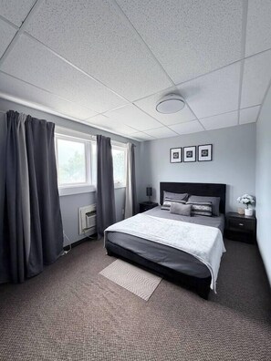 A bedroom with a bed, a nightstand, a lamp, a window with curtains, and a radiator.