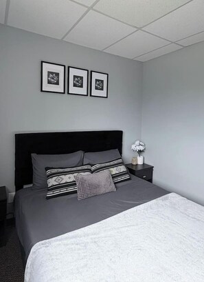 A neatly made bed with a dark headboard, a bedside table with a white vase, and three framed pictures on the wall. - Private 2BR Suite Near Downtown & Hospital (Regina)