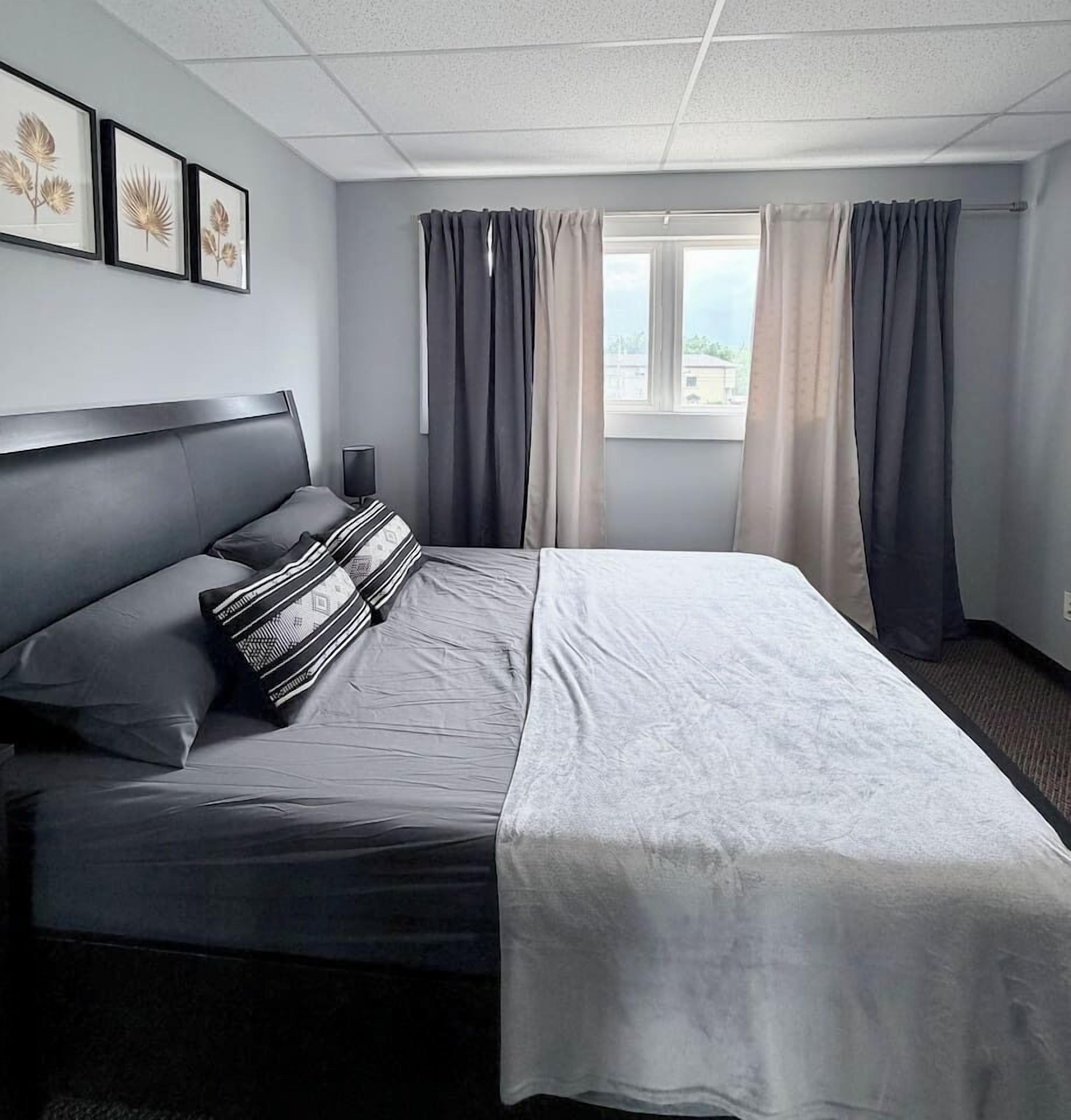 A hotel room with a bed, a bedside table, a window with curtains, and framed artwork on the wall.