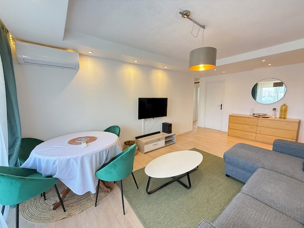A modern living room with a flat-screen TV, a sofa, a dining table with chairs, and a small coffee table.