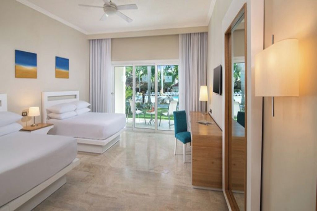 Quarto de hotel com duas camas, uma escrivaninha, uma cadeira e vista para a piscina e área externa.