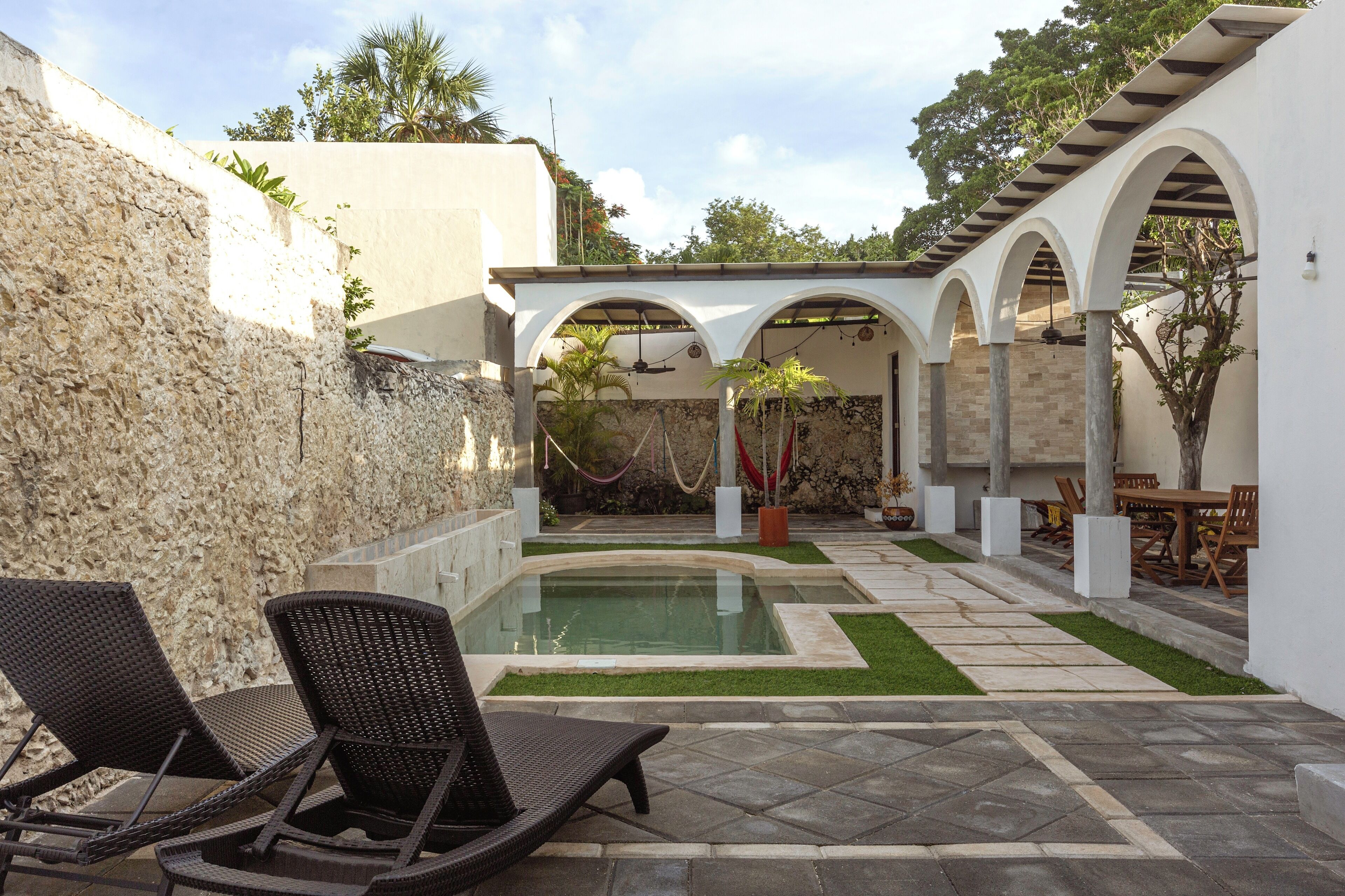 A pool area with lounge chairs, a pergola, and a stone wall.