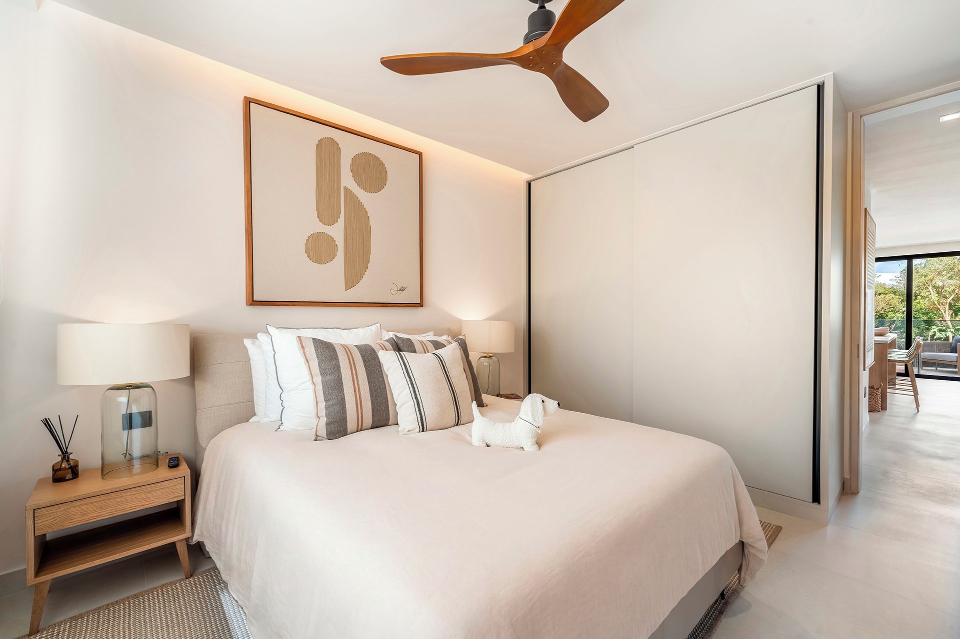 Une chambre à coucher moderne avec un grand lit, un ventilateur de plafond, une œuvre d’art abstraite encadrée et une vue sur l’extérieur.