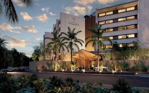 A modern hotel with a prominent entrance, a stone facade, and a well-lit outdoor area with palm trees. - Distrito Xcalacoco Beach (Playa del Carmen)