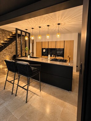A modern kitchen with a central island, bar stools, pendant lighting, and a stone staircase. - Exceptional Villa, Arcachon Bay, Infinity Pool (Le Teich)