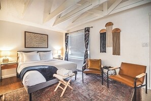 A hotel room with a bed, two chairs, a small table, a nightstand, a lamp, a window with blinds, and a wall-mounted artwork. - Cozy Studio Near Garden of the Gods - Fenced Yard (Colorado Springs)