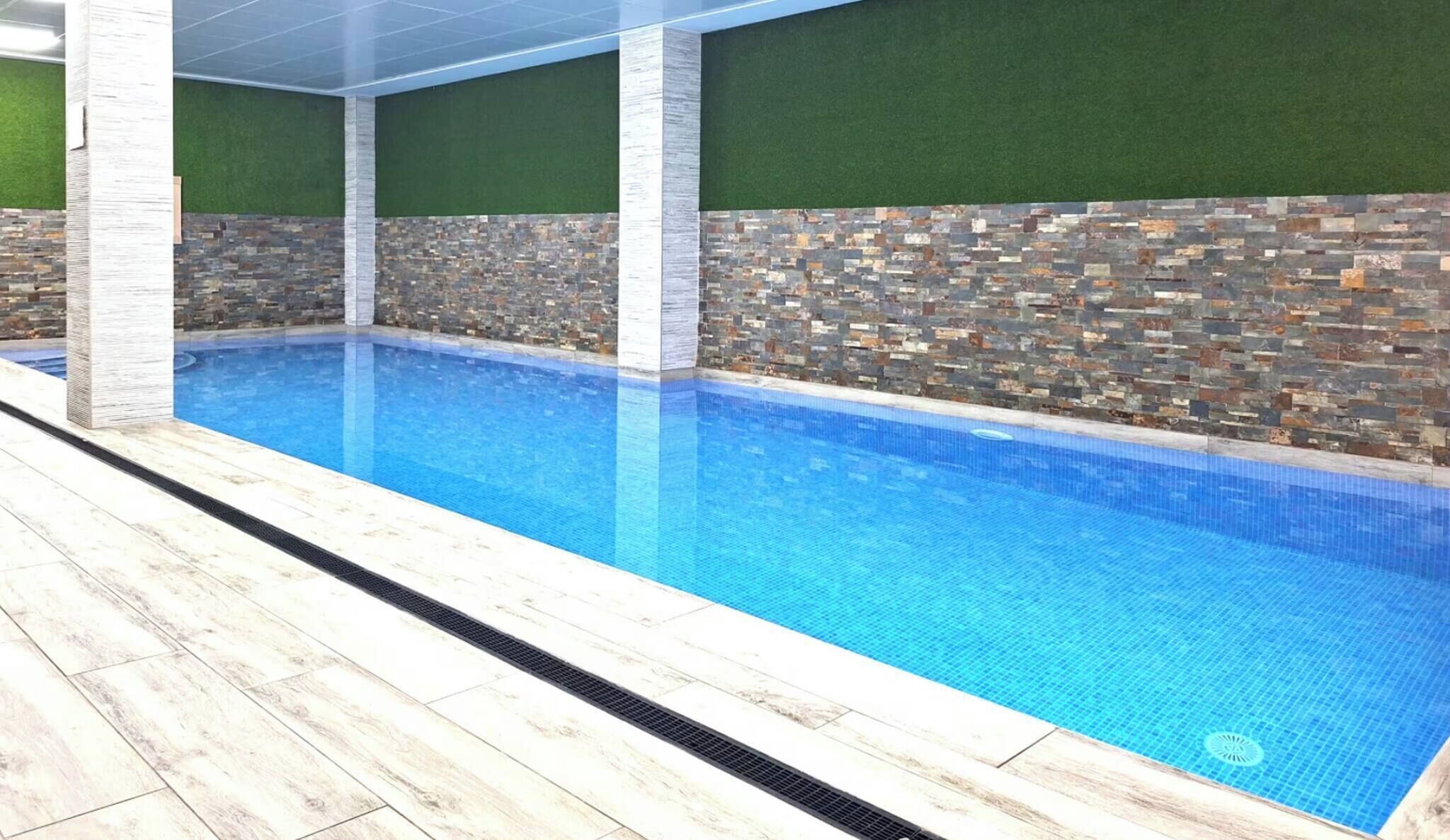 Une piscine dont le sol et les murs sont recouverts de carrelage, entourée d’un mur en pierre et d’une pelouse artificielle verte.