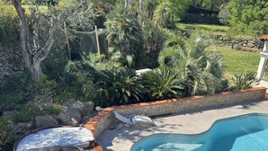 Une piscine avec un jacuzzi et des chaises longues, entourée d’un mur de pierre et d’une végétation luxuriante.