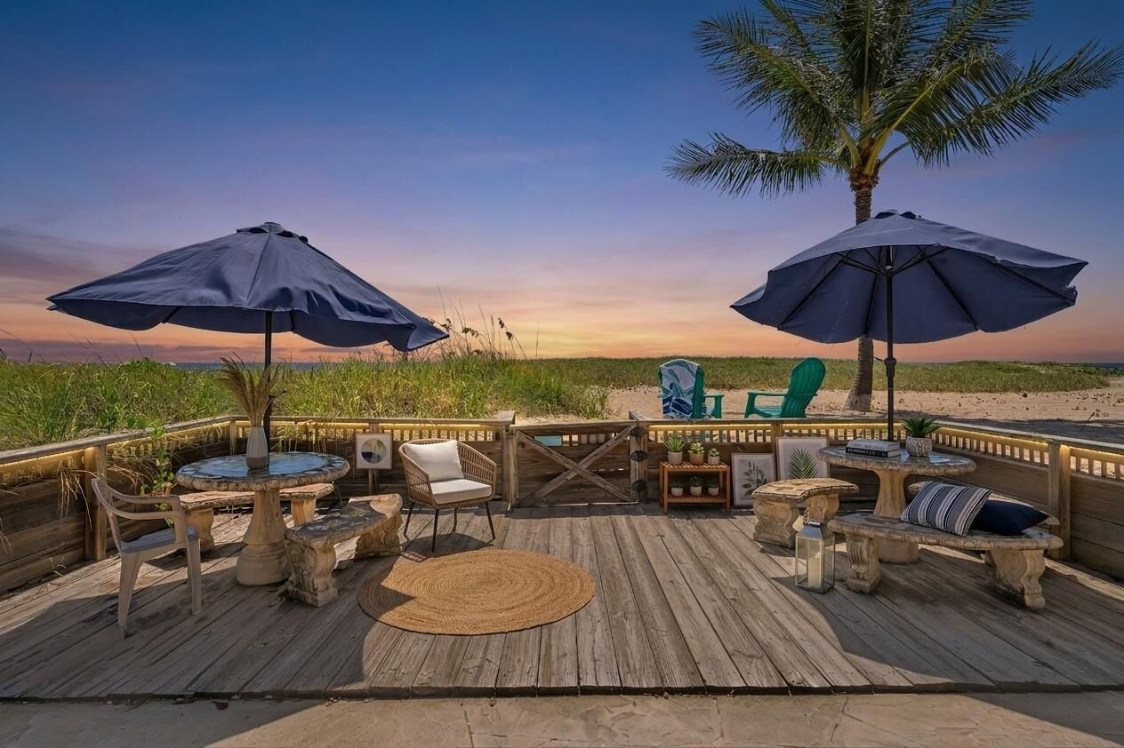 Une terrasse en bois, aménagée avec du mobilier d’extérieur, notamment des chaises et une table, donnant sur une plage bordée de palmiers.
