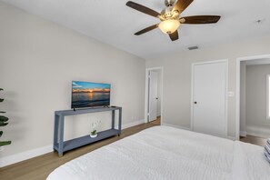 A bedroom with a bed, a ceiling fan, a console table with a plant, and a wall-mounted TV displaying a sunset scene. - LaPlaya Pompano Private Beach + Pool (Pompano Beach)