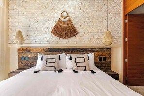 A bed with white bedding and two pillows with black tassels, a rustic wooden headboard, and a textured wall with a woven hanging decoration. - DÉJÁ VU PH 410 By SANDY POWERFUL (Tulum)