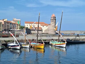 A harbor with boats docked, a stone tower, and buildings with red roofs. - The Birds of Paradise (VILLELONGUE DELS MONTS)