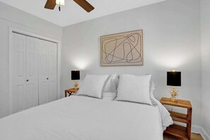A bedroom with a bed, two bedside tables with lamps, a ceiling fan, a door, and a wall-mounted abstract art piece.