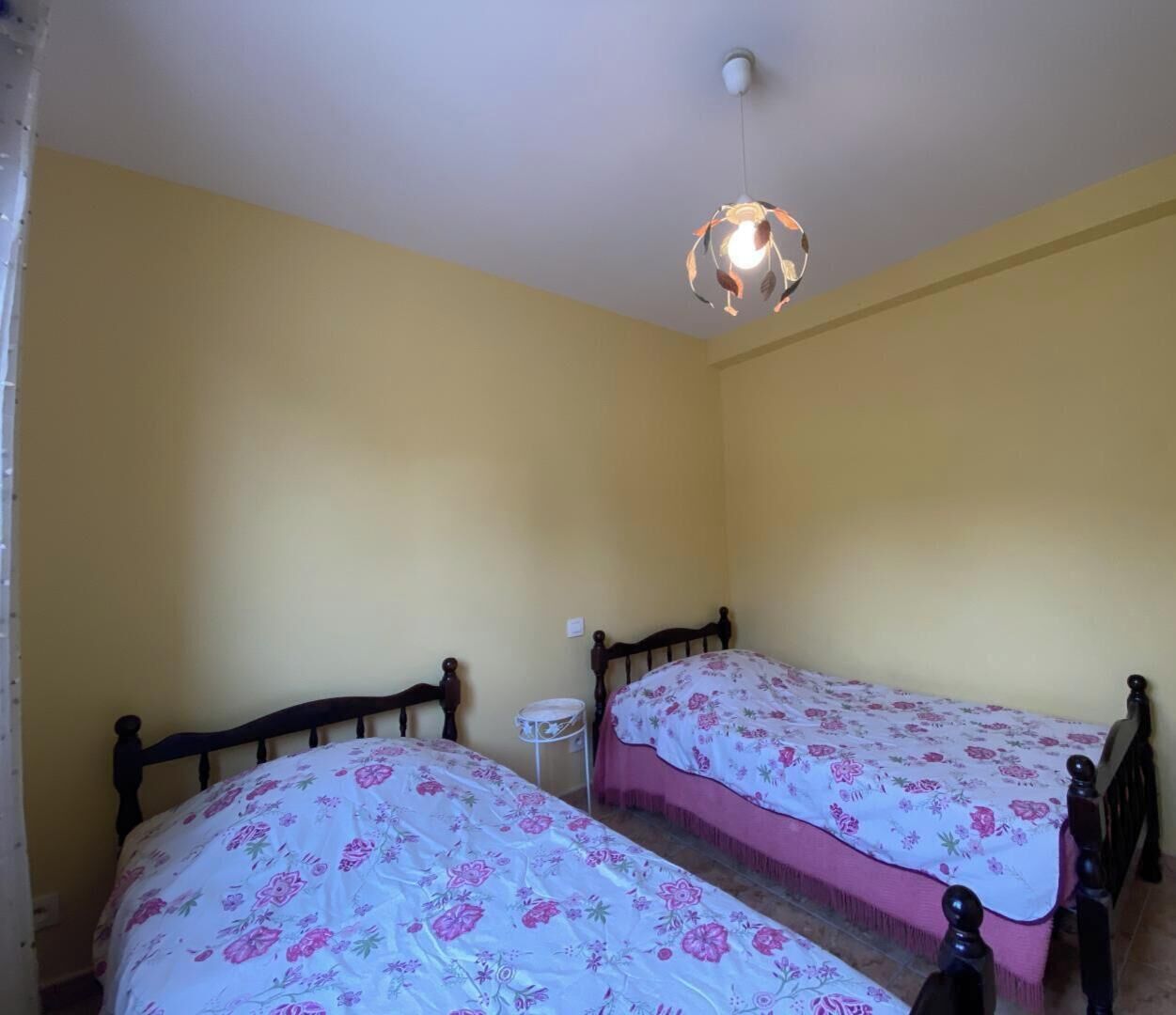 A room with two wooden beds, each with a floral bedspread, a small round table, and a hanging light fixture.