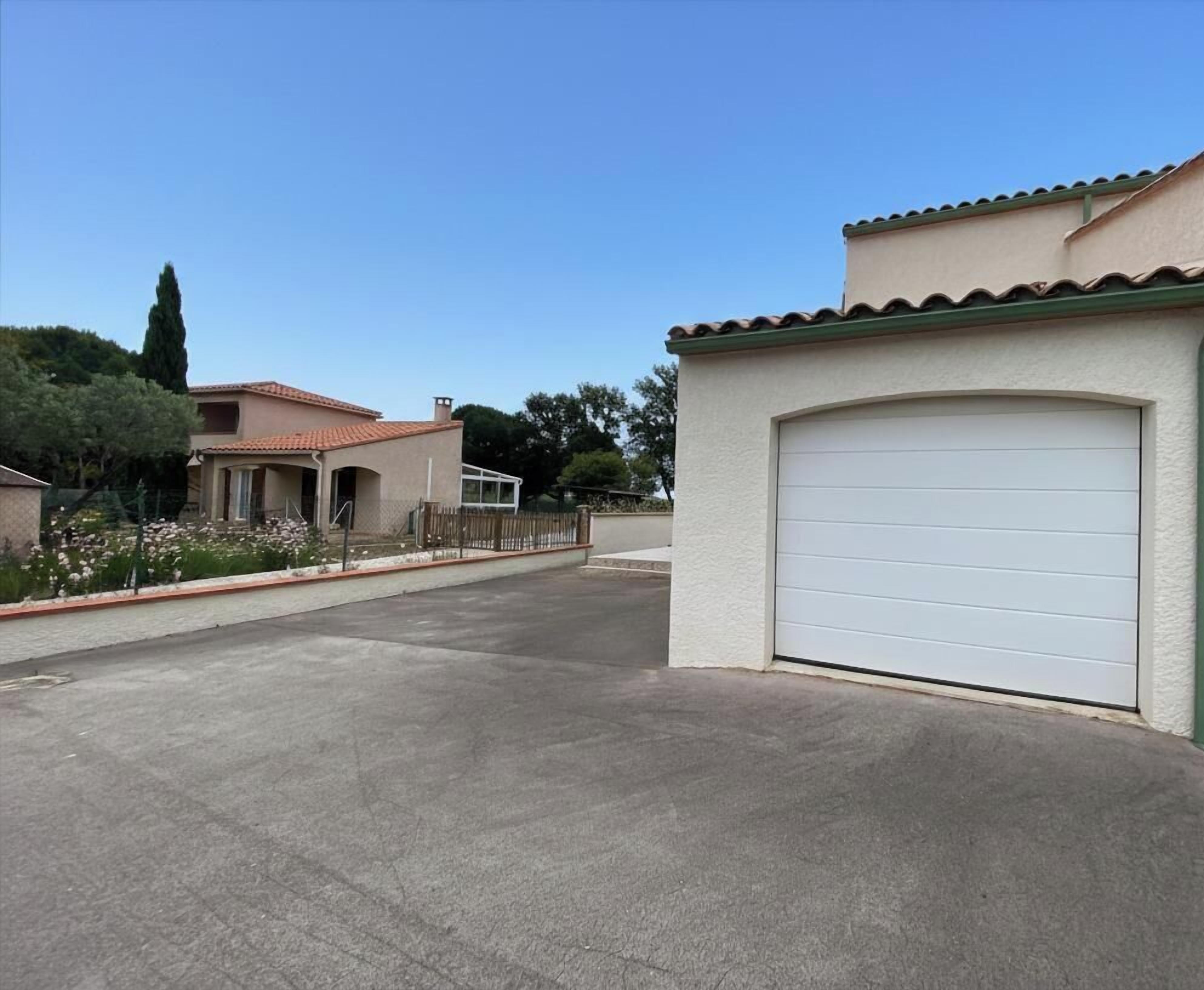 Uma entrada residencial com uma porta de garagem, uma casa com telhado ladrilhado e um jardim florido.