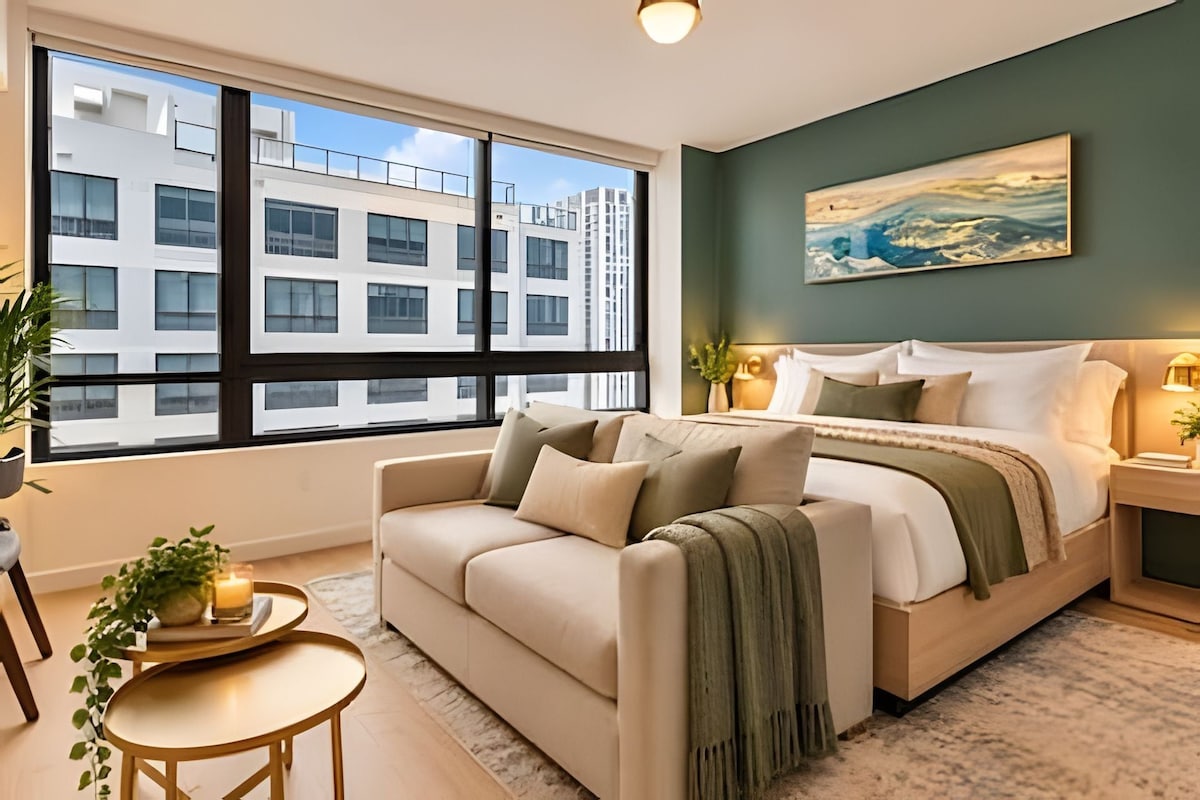 A modern bedroom with a large window, a bed, a sofa, a bedside table, and a painting on the wall.