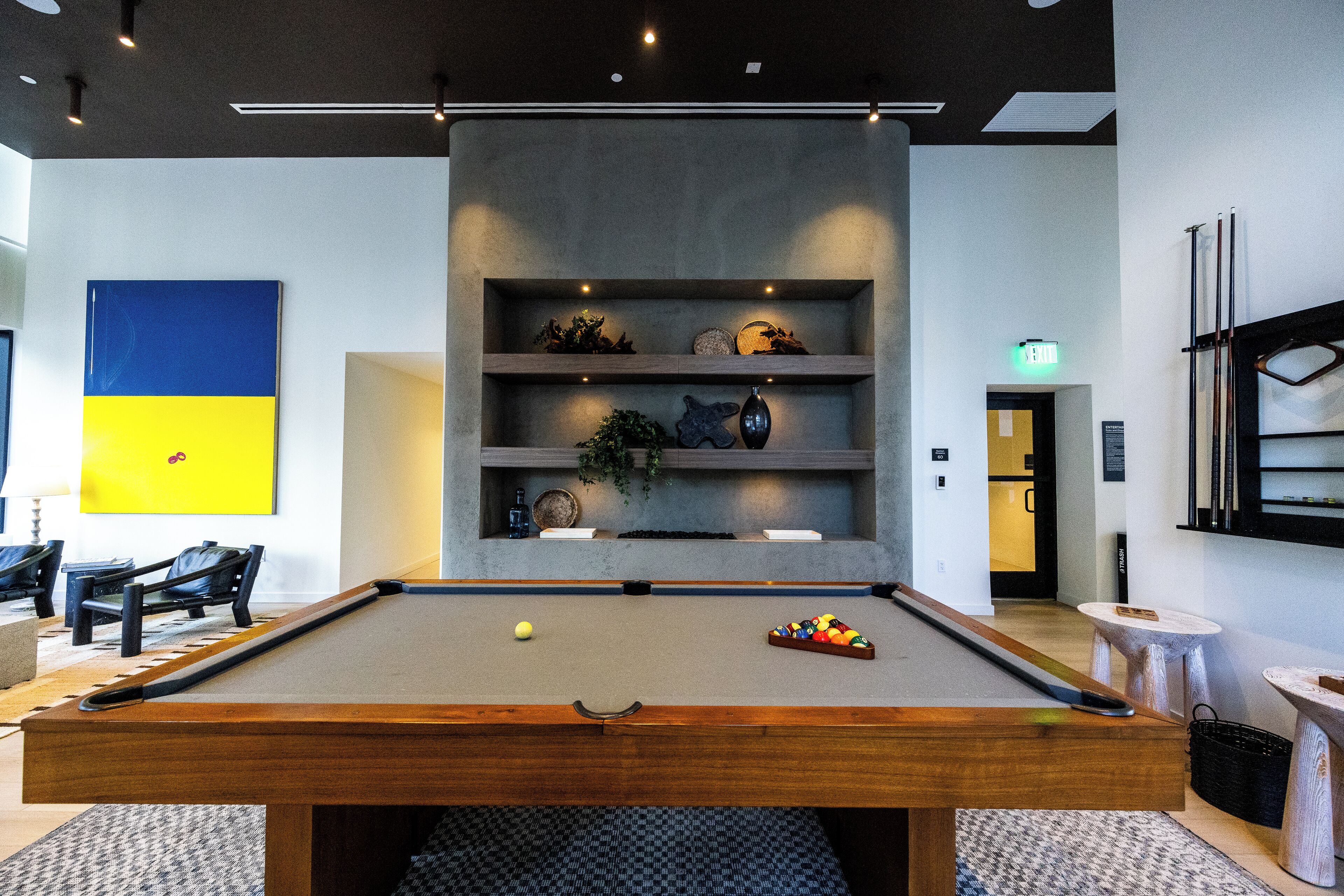A billiards table with balls and cues, a seating area with chairs, and a wall with a blue and yellow artwork.