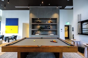 A billiards table with balls and cues, a seating area with chairs, and a wall with a blue and yellow artwork. - District 3704 (Miami)
