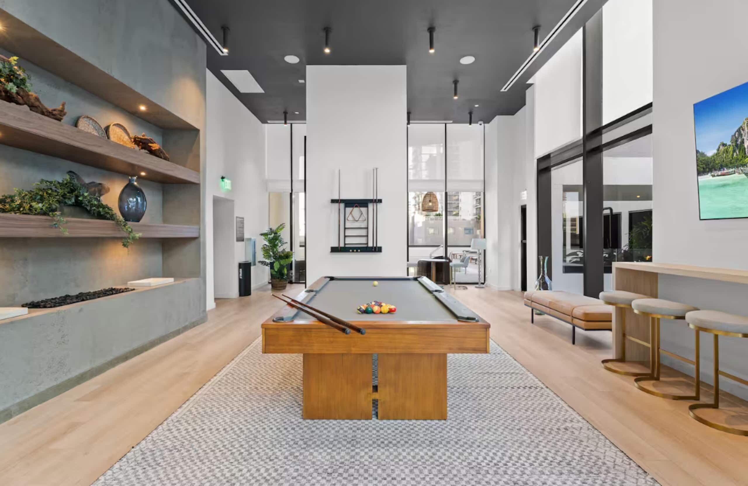 A modern indoor pool table with a wooden table and a patterned rug, surrounded by bar stools and a bench.