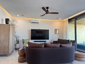 A modern living room with a flat-screen TV, a ceiling fan, and a large window. - Fabulous Penthouse with Ocean & Mountain Views (Puerto Vallarta)