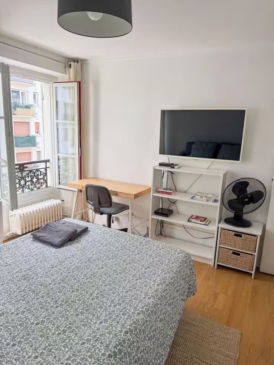 Une chambre à coucher comprenant un lit, un bureau, une chaise, un téléviseur, un radiateur et une fenêtre donnant sur des immeubles.