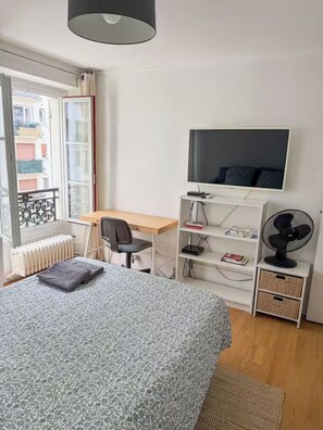 Een slaapkamer met een bed, bureau, stoel, televisie, radiator en raam met uitzicht op gebouwen.