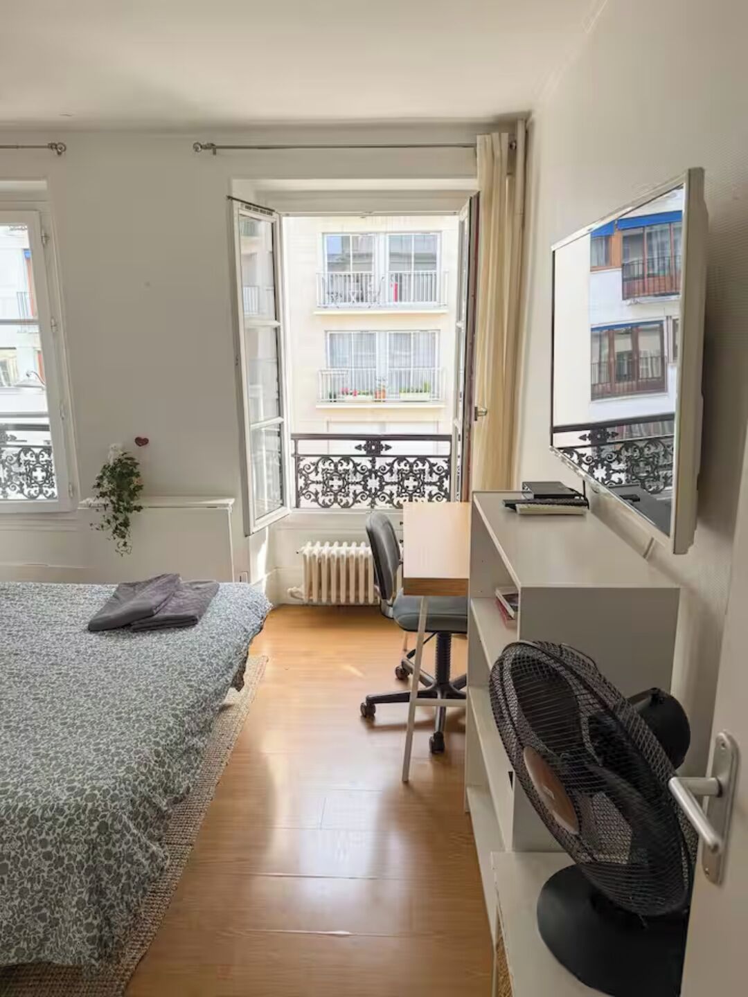 A bedroom with a bed, a desk, a chair, a radiator, and a window with a view of a balcony.
