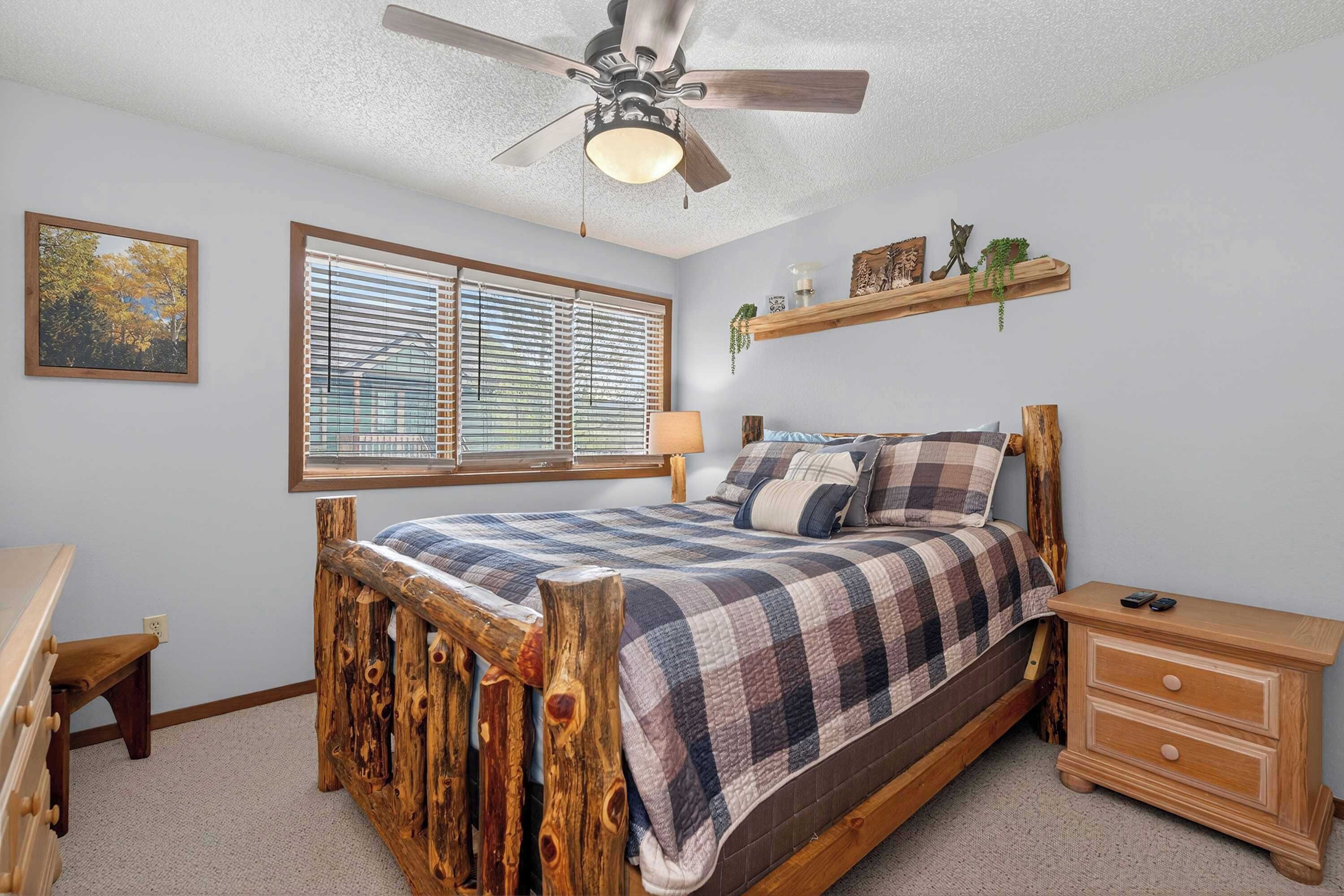 A bedroom with a wooden bed, a nightstand, a dresser, a ceiling fan, a window with blinds, and a wall-mounted shelf with decorative items.