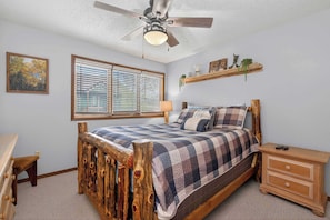 A bedroom with a wooden bed, a nightstand, a dresser, a ceiling fan, a window with blinds, and a wall-mounted shelf with decorative items. - WP Shuttle | Condo in Downtown Fraser (Fraser)