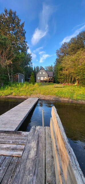 Puinen laituri, joka ulottuu tyynen vesialueen päälle, ja taustalla näkyy puilla ympäröity talo.