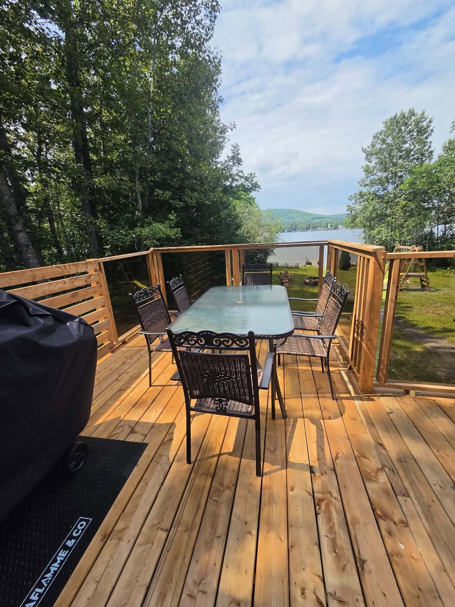 Une terrasse extérieure dotée d’une table et de chaises à plateau en verre, entourée de rampes en bois, et offrant une vue sur une étendue d’eau et sur la verdure.