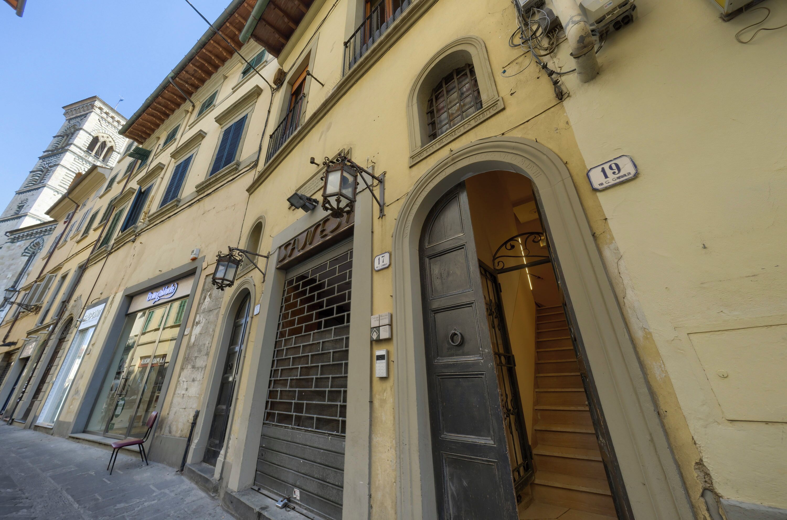 Una veduta di una strada con un edificio. Al piano terra si trova un negozio, una scala conduce a un piano superiore e un cartello con il numero 19 sulla parete.