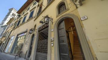 Una veduta di una strada con un edificio. Al piano terra si trova un negozio, una scala conduce a un piano superiore e un cartello con il numero 19 sulla parete.