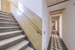 Eine Treppe mit Marmorstufen und Geländer, die zu einem Korridor mit Schachbrettfußboden und einem gemusterten Wandbild führt.