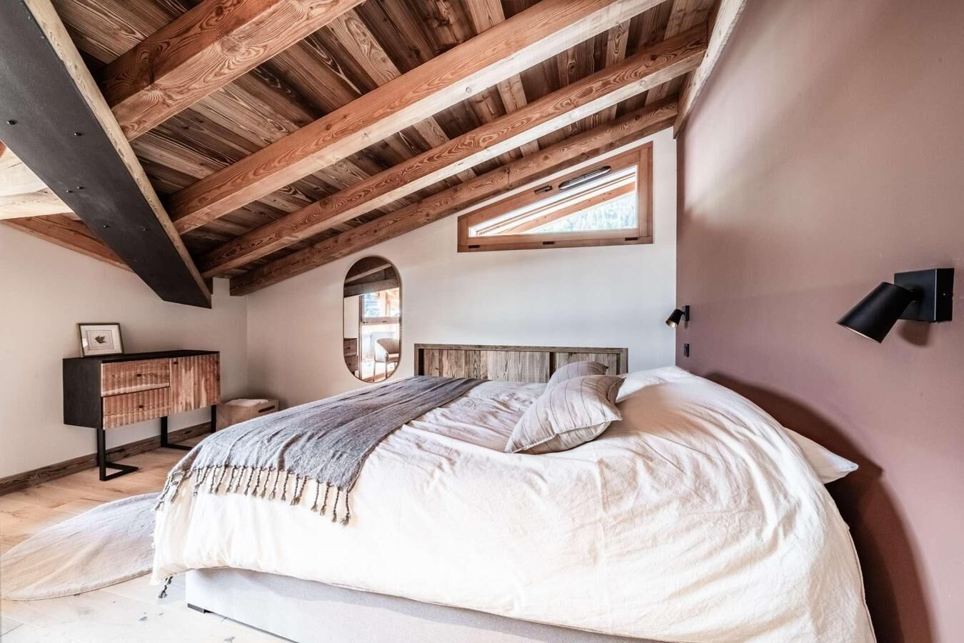 Une chambre avec un plafond en pente, un lit avec une literie blanche, une tête de lit en bois, une table de chevet et une lampe fixée au mur.